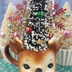 May include: A ceramic reindeer mug with a black and white Christmas tree on top, decorated with red, green, and gold ornaments. A red bow and bell adorn the top of the tree. Two small white Christmas trees are in the background.