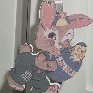 May include: A vintage-style Easter decoration featuring a winking brown bunny wearing green overalls. The bunny carries a blue egg with a chick inside. The decoration is made of paper and has a string for hanging.