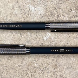 May include: Two black pens with silver accents. The top pen has the text "JOSEPH GORMAN" engraved on the side. The bottom pen has a series of dots and the text "HR" engraved on the side.