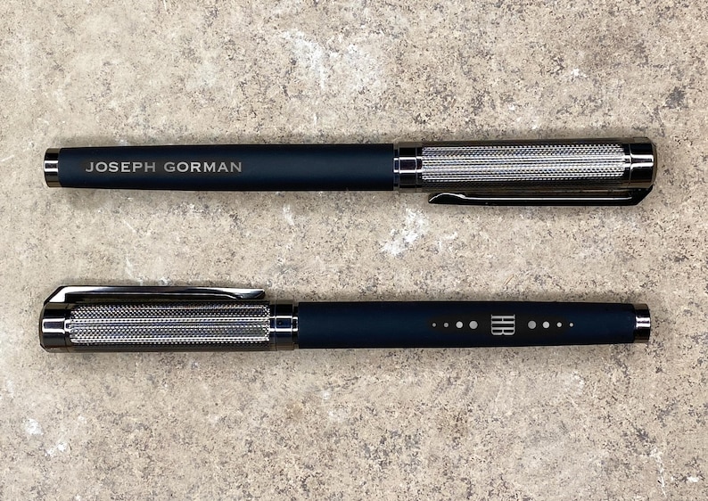 May include: Two black pens with silver accents. The top pen has the text "JOSEPH GORMAN" engraved on the side. The bottom pen has a series of dots and the text "HR" engraved on the side.