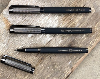 May include: Three black pens with silver accents and engraved text. The pens have a textured finish and are laid out on a wooden surface.