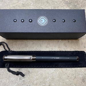 May include: A black pen with a silver band and the word "MC" engraved on it. The pen is in a black velvet pouch and a black box with a blue circle logo.