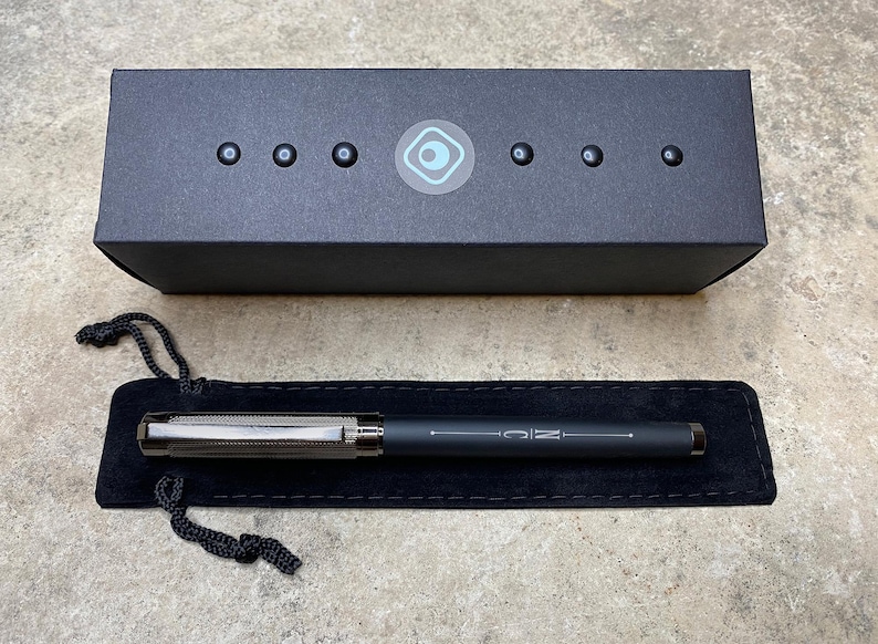 May include: A black pen with a silver band and the word "MC" engraved on it. The pen is in a black velvet pouch and a black box with a blue circle logo.