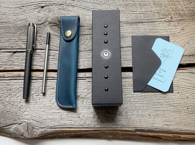 May include: A blue leather pen case, a silver pen, a silver mechanical pencil, a black box with a silver logo, and a blue card with the text "Welcome to the team, Emma!" in a black envelope.