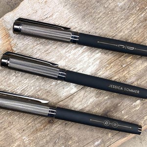 May include: Three black pens with silver accents and engraved names. The pens have a textured metal barrel and a black clip.