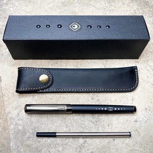 May include: A black leather pen case with a gold button closure, a black pen with silver accents and a silver pen with a black tip. The pen case and pens are on a gray textured surface.