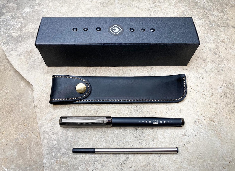 May include: A black leather pen case with a gold button closure, a black pen with silver accents and a silver pen with a black tip. The pen case and pens are on a gray textured surface.