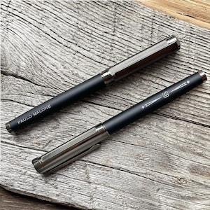 May include: Two black pens with silver accents. The pen on the left has the words "PAULO MALONE" engraved on it. The pen on the right has the letters "SB" engraved on it.