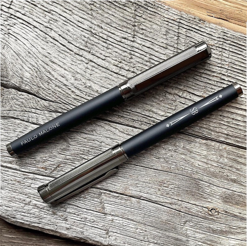 May include: Two black pens with silver accents. The pen on the left has the words "PAULO MALONE" engraved on it. The pen on the right has the letters "SB" engraved on it.