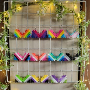Puede incluir: Una muestra de 10 pares de pendientes hechos a mano con la bandera del arcoíris del orgullo. Cada par está hecho de cuentas de perler de colores y presenta un diseño diferente de la bandera del orgullo. Los pendientes se muestran en una rejilla blanca.