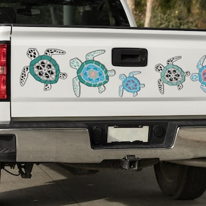 May include: White truck tailgate with five colorful turtle decals. The turtles have teal, blue, and pink accents. The truck has a chrome bumper and red taillights. The decals are arranged horizontally.