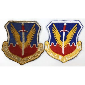 May include: Two embroidered patches with a shield design. The patches feature a blue background with a gold sword and wings, a red base, and the text "TACTICAL AIR COMMAND". The patches are edged in gold.