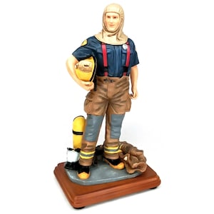 May include: A figurine of a firefighter in a navy blue shirt with red suspenders, brown pants, and a tan hood. The firefighter is holding a yellow helmet. A yellow oxygen tank and gloves are at the base. The figurine is on a wooden base.