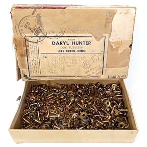 May include: An open cardboard box filled with various brass and copper rivets and fasteners. The box has a label that reads "DARYL HUNTER DOG SUPPLIES LEES CREEK, OHIO". The box is labeled "THIRD CLASS".