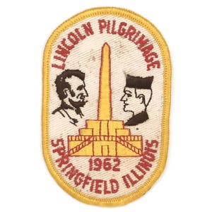May include: An oval patch with a yellow border features the text "Lincoln Pilgrimage Springfield Illinois 1962." The patch includes illustrations of Abraham Lincoln and another man, along with a yellow and orange monument.