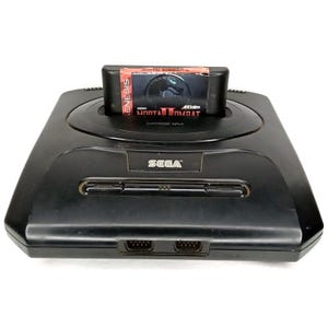 May include: A black Sega Genesis console with a "Mortal Kombat II" game cartridge inserted. The cartridge is red, black, and white. The console has the word "SEGA" printed on the front.