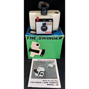 May include: Vintage Polaroid Swinger camera with a cream-colored body and a black lens. The camera is on top of a green box with the words "THE SWINGER" and a diagram. A manual titled "How to use the POLAROID LAND CAMERA MODEL 20" is in front.