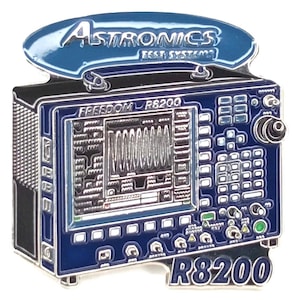 May include: A blue and silver pin featuring a detailed illustration of a piece of electronic test equipment. The pin has the text "Astronics Test Systems" at the top and "Freedom R8200" and "R8200" on the front.