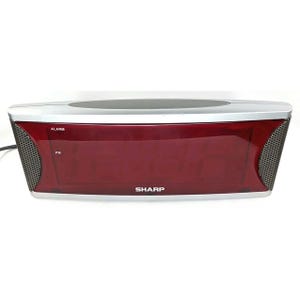May include: A rectangular digital alarm clock with a red display and silver accents. The clock has a mesh speaker on each side and the brand name "SHARP" is visible. The display shows "ALARM" and "PM".