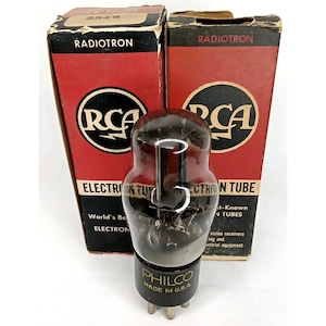 May include: A vintage vacuum tube with a dark glass bulb and a base marked "PHILCO MADE IN U.S.A." is displayed in front of two red and black RCA Radiotron boxes. The boxes feature the RCA logo and text.