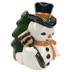 May include: A glossy ceramic snowman figurine wearing a black top hat with holly berries, an orange scarf, and three black buttons. The snowman holds a brown broom and stands in front of a dark green pine tree, all finished with a smooth glaze.
