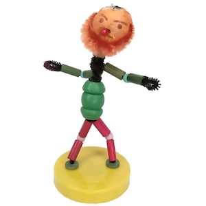 May include: A whimsical pipe cleaner figure with a red beard, green torso, and red legs. The figure is standing on a yellow base.