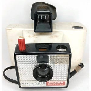 May include: Vintage Polaroid Swinger Model 20 Land Camera. The camera is black with a silver lens and a red button. The viewfinder is black and sits atop a white camera body. A black strap is attached.
