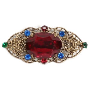 May include: Vintage brooch with a large, faceted red gemstone as the focal point. The brooch features a filigree design in gold-tone metal, with smaller blue, red, and green accent stones. The brooch has an oval shape.