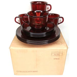 May include: A set of 12-piece ruby red glassware, including cups and saucers, stacked on top of a cardboard box. The cups have handles and the plates are round. The box has text that reads "RUBIS" and "12 PIECES".