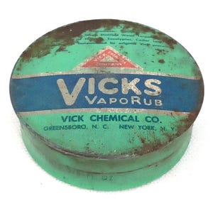 May include: Vintage Vicks VapoRub tin. The round tin is green with a blue band across the center. The text "VICKS VAPORUB" is in silver letters. The tin also includes the text "VICK CHEMICAL CO. GREENSBORO, N.C. NEW YORK, N.Y."