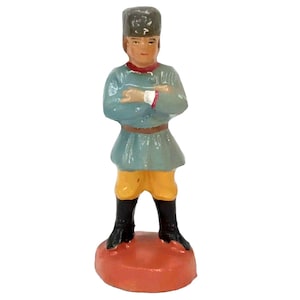 May include: A small figurine of a person wearing a blue and yellow outfit and a black fur hat. The figurine is standing on a red base.
