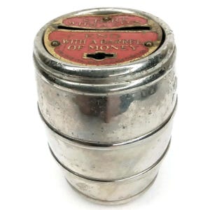 May include: A vintage silver-colored metal coin bank shaped like a barrel. The top has a red insert with the text "START WITH A COIN" and "END WITH A BARREL OF MONEY". The bank has a slot for coins and a keyhole.