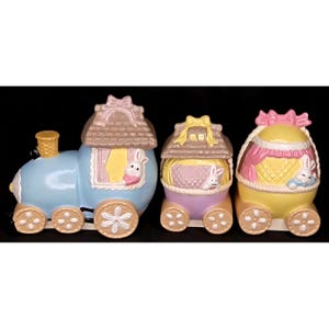 May include: A pastel-colored Easter train set with three carriages. The train engine is light blue, and the carriages are purple and yellow. Each carriage has a bunny peering out of a window. The train wheels are decorated with flower shapes.
