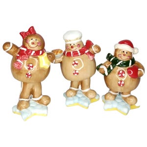 May include: Three ceramic gingerbread figures with festive holiday attire. Each figure has a brown body, white icing accents, and a candy button. One wears a chef's hat, another a Santa hat, and the third a red bow. They stand on blue star-shaped bases.