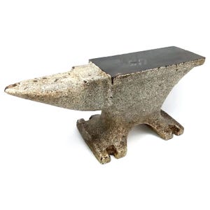 May include: A heavy metal anvil featuring a smooth, dark, flat top surface for striking and a textured, weathered, light gray body. The anvil has a pointed horn on one side, a wide rectangular face, and a sturdy base with four feet.