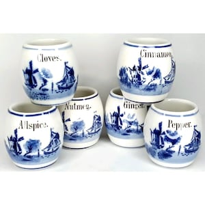 May include: Set of six white ceramic spice jars with blue windmill and sailboat illustrations. Each jar is labeled with a spice name: Cloves, Cinnamon, Allspice, Nutmeg, Ginger, and Pepper. The jars have a classic, vintage design.