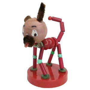 May include: A red and green wooden toy dog with a brown yarn tail and a painted face. The dog is on a red base.