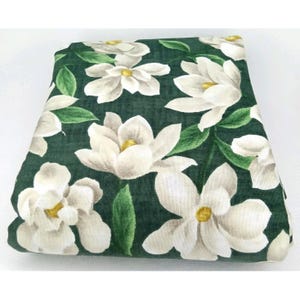 May include: Folded fabric with a dark green background and a pattern of large white magnolia flowers with yellow centers and green leaves. The fabric is folded neatly, showcasing the floral design.