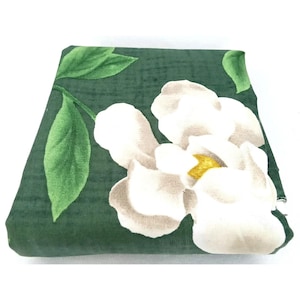 May include: Folded green fabric with a large white magnolia flower print. The flower has a yellow center and is surrounded by green leaves. The fabric is folded neatly, showcasing the floral pattern.
