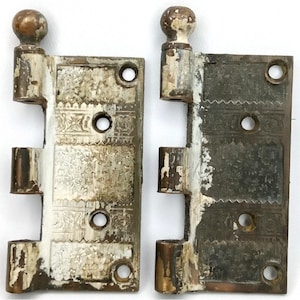 May include: Two antique door hinges with decorative floral and geometric patterns. The hinges are made of metal with a distressed, aged appearance, featuring peeling white paint and exposed bronze. Each hinge has a rounded top.