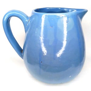 May include: A light blue ceramic pitcher with a curved handle and a spout. The pitcher has a glossy finish and a rounded body. The handle is attached to the side of the pitcher, and the spout is located at the top. The pitcher is likely used for serving beverages.
