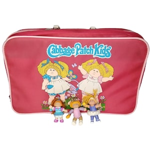 May include: A rectangular pink suitcase featuring the Cabbage Patch Kids logo and a graphic of two blonde dolls in white dresses. Three small Cabbage Patch Kids figurines are positioned in front of the suitcase, wearing colorful outfits.