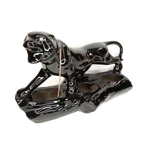 May include: A glossy black ceramic panther figurine with a gold chain collar. The panther is posed atop a black, textured base with three openings. The figurine is a decorative item.