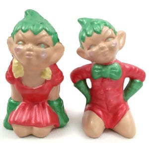 May include: Two ceramic figurines of elves. One elf is wearing a red dress with green accents and yellow shoulder details. The other elf is kneeling, wearing a red bodysuit with a green bow tie and green gloves. Both have green hair.