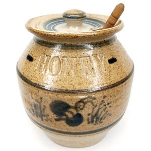May include: A tan ceramic honey pot with a lid and a wooden honey dipper. The word "HONEY" is carved into the front of the pot. The pot has blue painted accents and a floral design around the base.