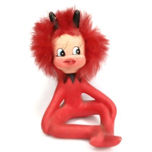 May include: A vintage figurine of a seated devil with a red body and fluffy red hair. The devil has a painted face with blue eyes, freckles, and a red mouth. Black horns and a black collar complete the design. The figurine is on a white background.