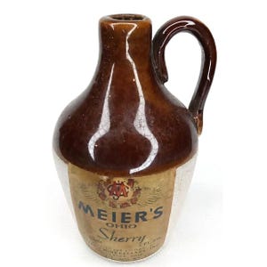 May include: A brown and white ceramic jug with a handle. The jug has a label that reads "Meier's Ohio Sherry". The jug is likely vintage and has a glossy finish.