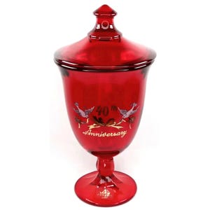 May include: A ruby red glass candy dish with a lid. The dish has a pedestal base and a flared bowl shape. The front of the dish features the text "40th Anniversary" with two birds. A decorative piece for a special occasion.