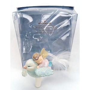 May include: A Hallmark Keepsake Ornament featuring a small angel with a pink outfit and a floral crown resting on a light blue bird with glittery wings. The ornament is packaged in a clear plastic box with the Hallmark Keepsake logo printed on it.