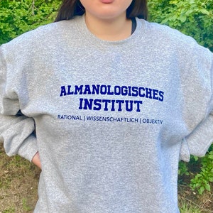 Könnte beinhalten: Grauer Pullover mit dem Text "ALMANOLOGISCHES INSTITUT" in blauer Schrift und den Worten "RATIONAL | WISSENSCHAFTLICH | OBJEKTIV" in kleinerer blauer Schrift darunter.